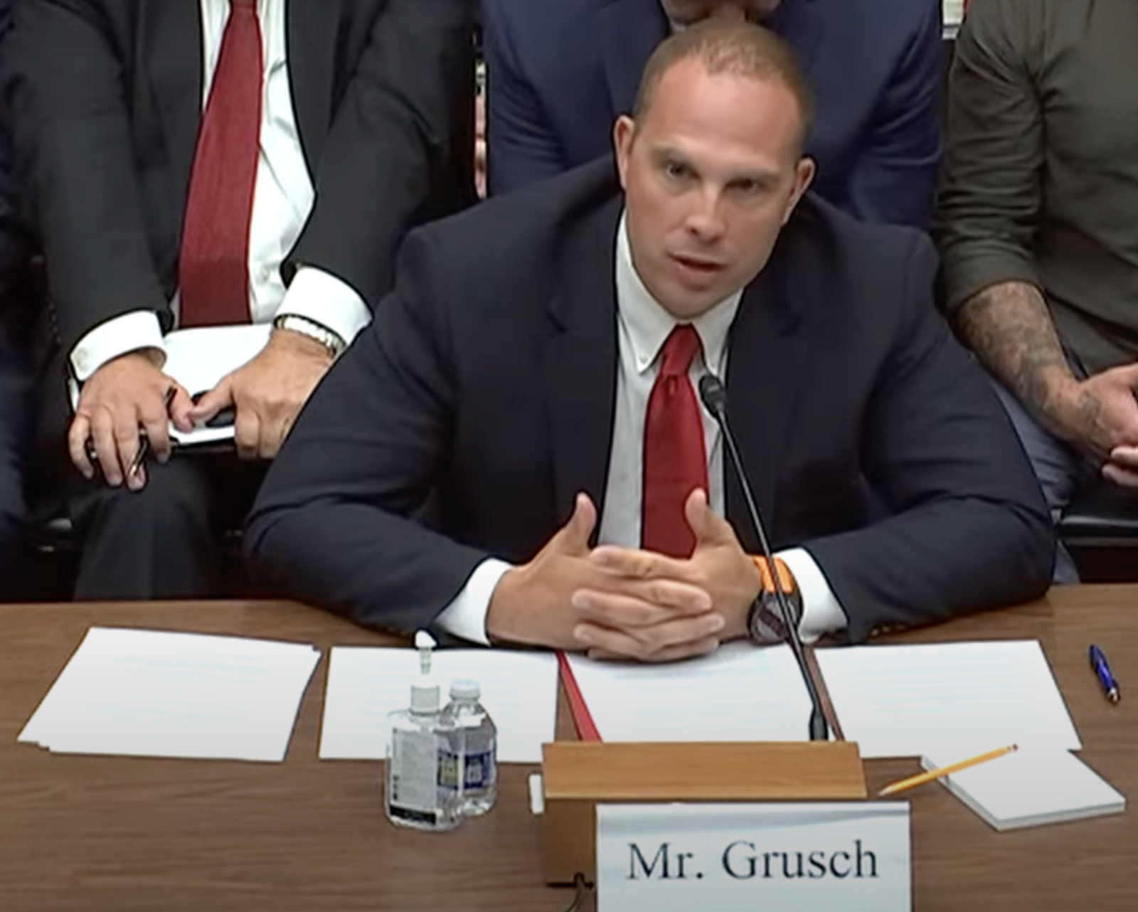 David Grusch, in a dark suit, seated at a witness table speaking into a microphone during a congressional hearing