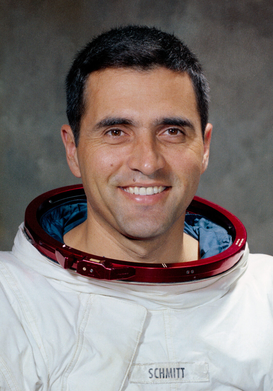 NASA portrait of astronaut Harrison H. Schmitt in a white Apollo pressure suit, helmet held at his side, against a lunar-surface backdrop.